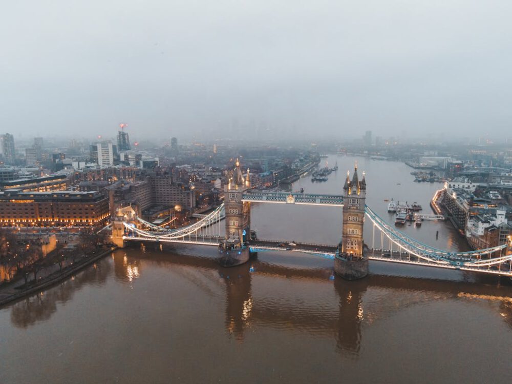 Drone view of London city located in England with illuminated Tower Bridge on River Thames near buildings in misty weather under gray cloudy sky in daylight